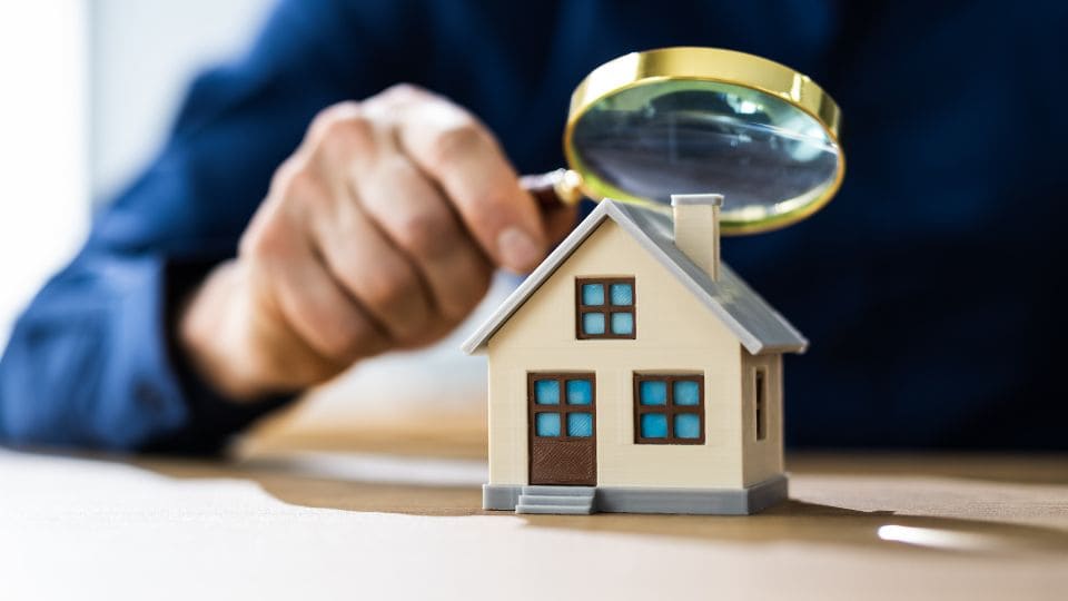 Person using a magnifying glass to closely examine a model house, representing real estate investment decisions on where to buy and sell amidst climate change